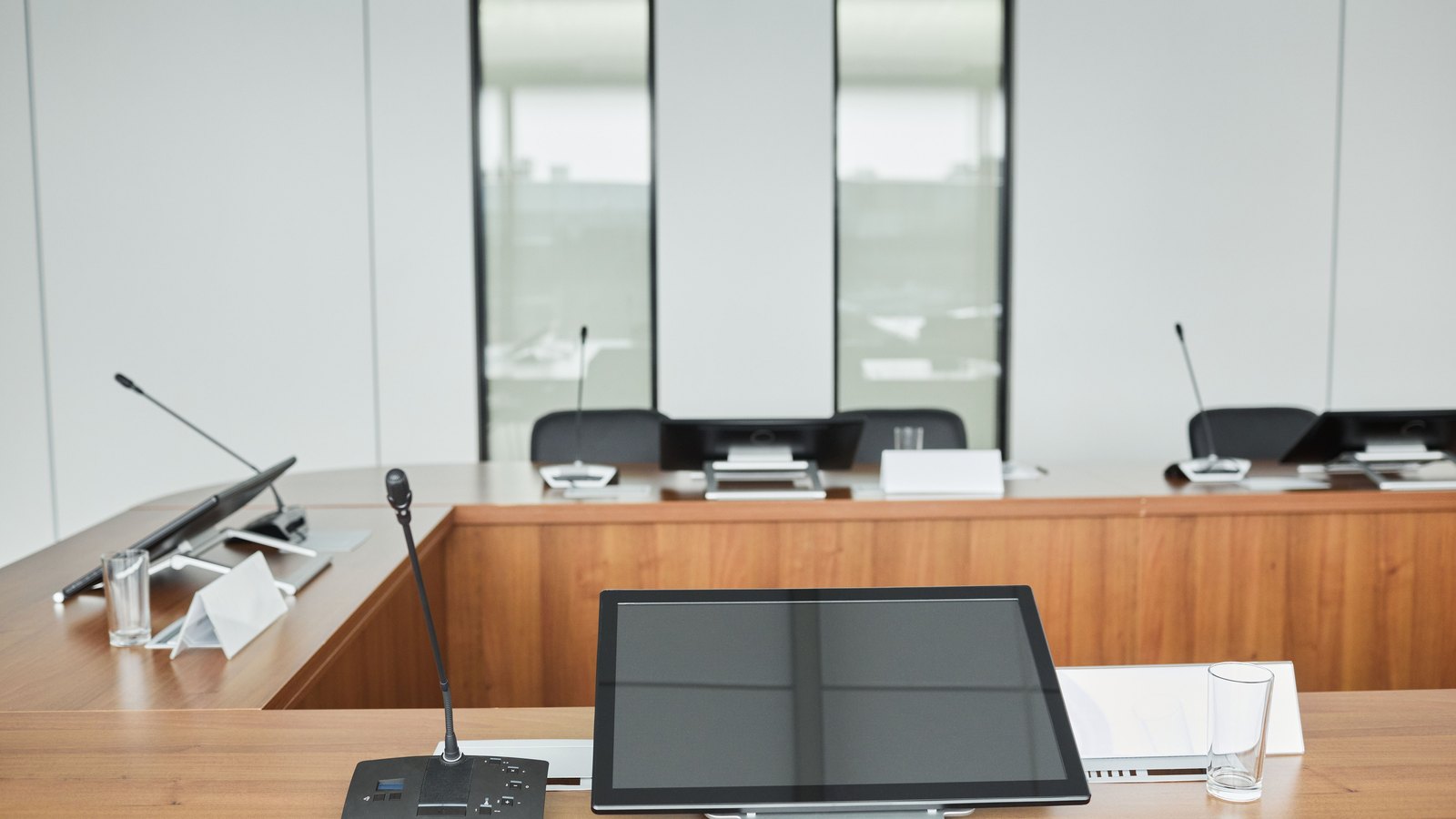 An empty modern council room seen across a light wood dais. A gooseneck microphone and a blank flat screen monitor sit at an unoccupied place in the foreground, with a water glass and a folded paper card beside them, and three more empty places with their own microphones curve away behind. The chairs are black and all of them are empty. There is no person in the frame and no legible lettering, signage, seal or place name of any kind. Illustration of the kind of municipal meeting room where a council sits, not a photograph of this event.