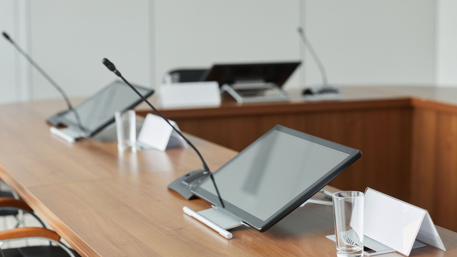 An empty meeting table of pale wood set for a formal session, with two gooseneck microphones, two flat monitor screens switched off, drinking glasses and folded white name cards that carry no visible lettering. A wooden bench and a further microphone stand behind. Nobody is in the frame and nothing identifies a place. It is a stock photograph of an unoccupied council style dais and not a photograph of the Cape Coral council chamber.