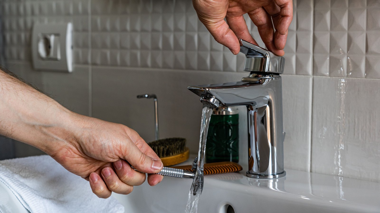 Cropped hands hold an electric shaver under water flowing from a chrome bathroom faucet, with no face or legible text visible. A stock photograph, not a photograph of this event.