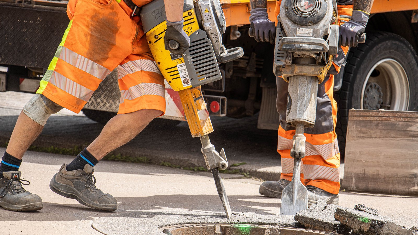 Two road workers in orange high visibility trousers breaking a section of asphalt around an open manhole with handheld pneumatic tools, beside a work truck. Both are framed from the waist down so no face is in view, and no signage, logo, plate or location identifies the scene. A generic illustration of road repair work, not a photograph of this event.