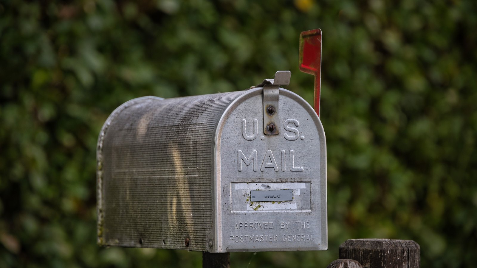 A weathered aluminium roadside mailbox of the standard domestic pattern, its red flag raised, mounted on a post beside a wooden fence rail against a wall of blurred green foliage. Nothing in the frame carries an address, a house number, a plate or any mark identifying a person or a location. A generic illustration of household mail collection, not a photograph of this event.