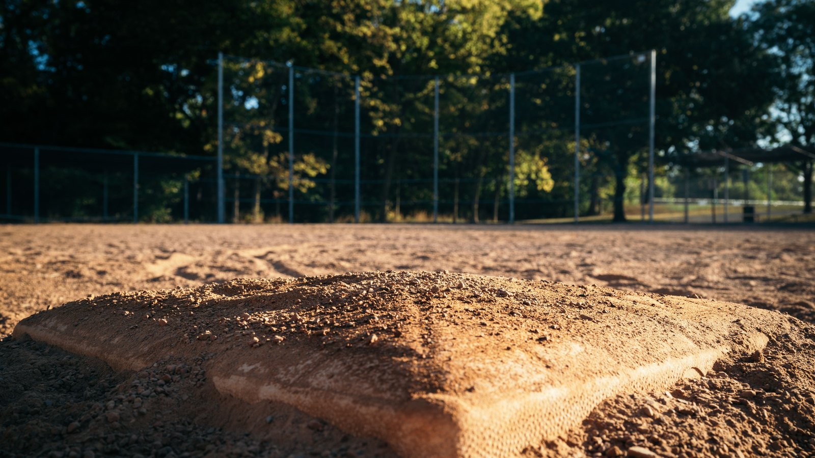 A low-angle photograph taken at the level of a baseball base, which fills the foreground and is scattered with loose dirt and small stones. Behind it the infield runs flat and empty to a chain link backstop, with mature deciduous trees in low evening sun beyond and a small open shelter at the right edge. No people, no lettering, no signage and no place can be identified from it. A generic illustration of a ball field, not a photograph of the Northwest Softball Complex or any Cape Coral park.