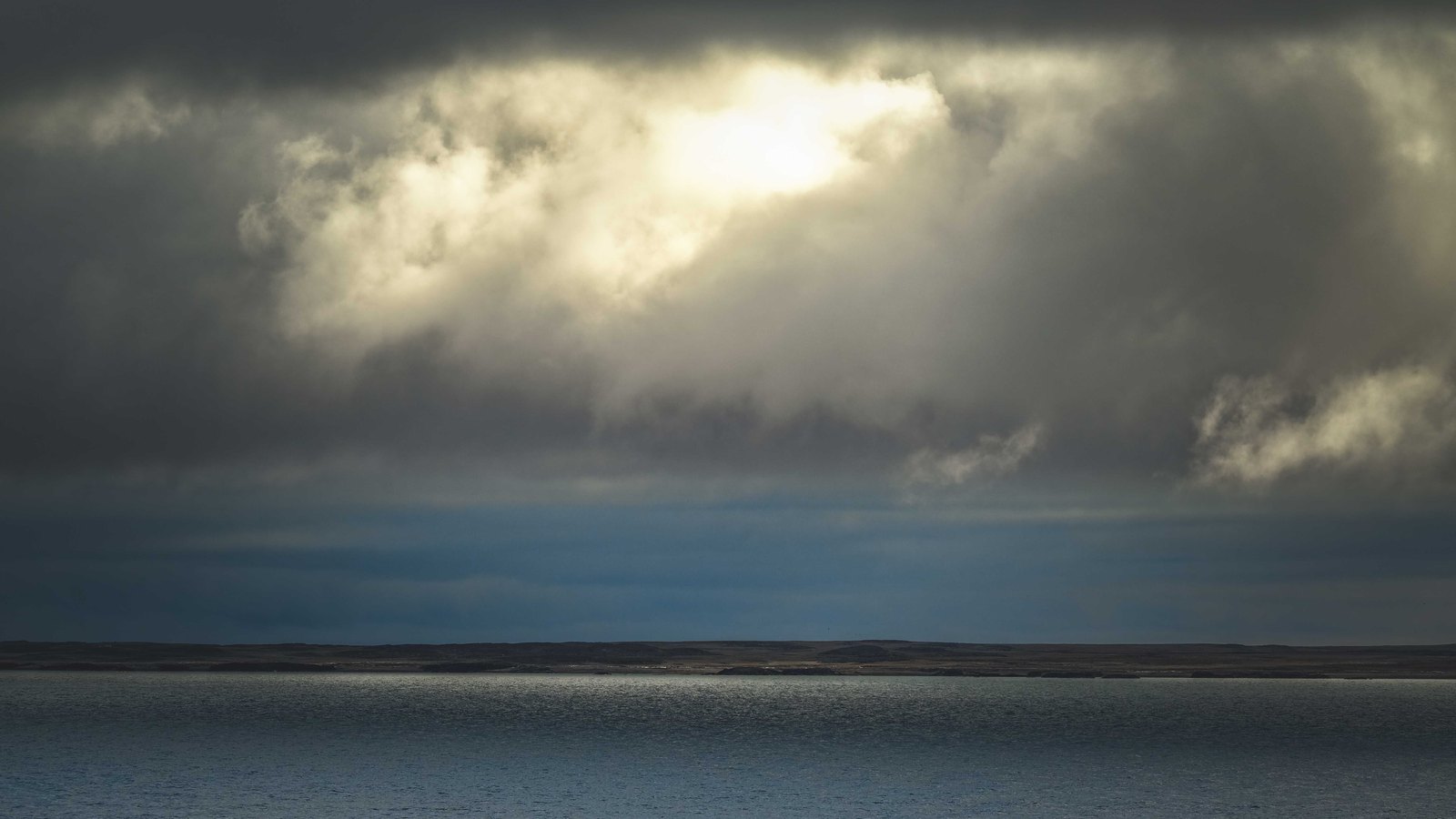 A heavy grey storm cloud spread across the whole sky above a wide stretch of open water, broken in the upper middle where sunlight burns through a gap and lights the cloud from behind. A low, flat, bare shoreline runs along the horizon. No signage, lettering or place name is in the frame. A generic illustration, not a photograph of Charlotte Harbour or Cape Coral: the distant shore is treeless and bare rather than the mangrove and development of this coast, and the light is daytime rather than the early evening this storm sat over the county.