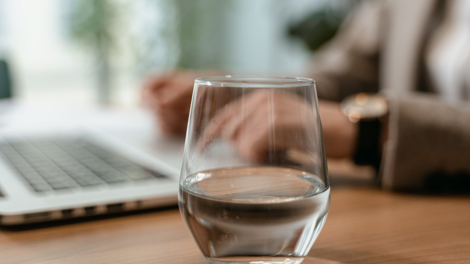 A clear glass of water in focus on a desk, with a blurred person at a laptop behind it, not a photograph of this event.