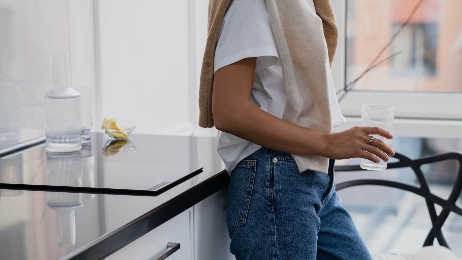A person in a white t-shirt and jeans, cropped at the neck so no face is shown, leaning against a kitchen counter and holding a plain glass of clear water. A carafe and a second empty glass stand on the black counter behind them. No face, no signage, no legible text, no address and no identifiable place appears in the frame. A stock photograph, not a photograph of this event.