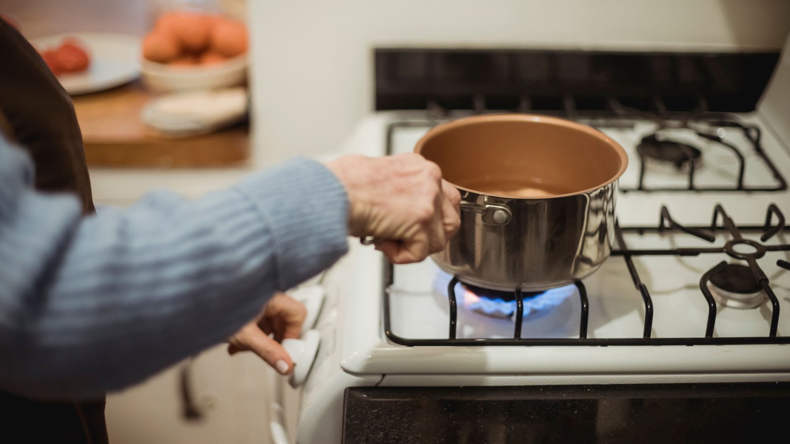 A photograph of a person in a blue jumper setting a stainless steel saucepan onto a lit gas ring on a white cooker, one hand on the pan handle and the other turning the control knob. A blue flame is visible under the pan. A kitchen counter is out of focus behind. No lettering or brand mark is in the frame.