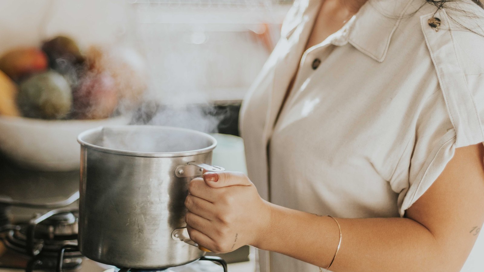 A steaming stainless steel pot on a gas hob, a hand resting on the handle, the person cropped above the chin so no face is visible. No text, no address, no vehicle and no agency marking appears in the frame. A stock photograph, not a photograph of this event.