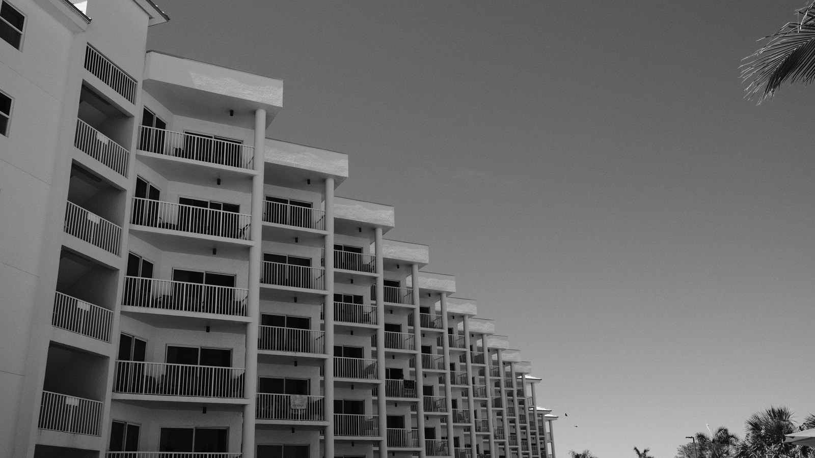 A black and white photograph looking up at the long side of a condominium building, its stacked balconies with slatted railings receding in a row against a blank pale sky, with palm fronds entering the frame at the right. There are no people, no text, no signage, no brand marks and no landmark by which any place could be identified. A generic illustration of condominium living, not a photograph of Cape Coral, of Southeast 5th Place, or of any building inside the boil water area. Its weakness is stated rather than glossed: the building is a tall resort style block, while the communities named in this item are smaller Cape Coral condominium developments, and the picture is monochrome, so it should not be read as showing what any of them look like.