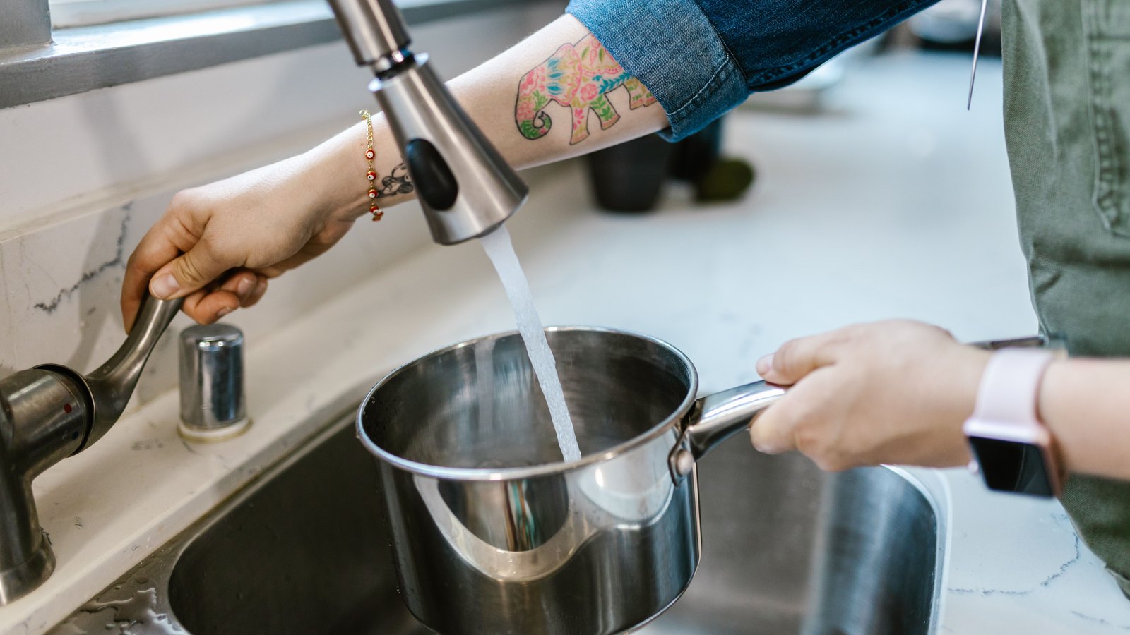 A photograph of two people at a kitchen sink filling a stainless steel saucepan from a chrome pull down tap. One hand, with a colourful elephant tattoo on the forearm, holds the tap lever open while another hand steadies the pan by its handle under the running water. A white worktop and a stainless steel sink surround them. No signage or legible text is in the frame. A generic illustration, not a photograph of Cape Coral.
