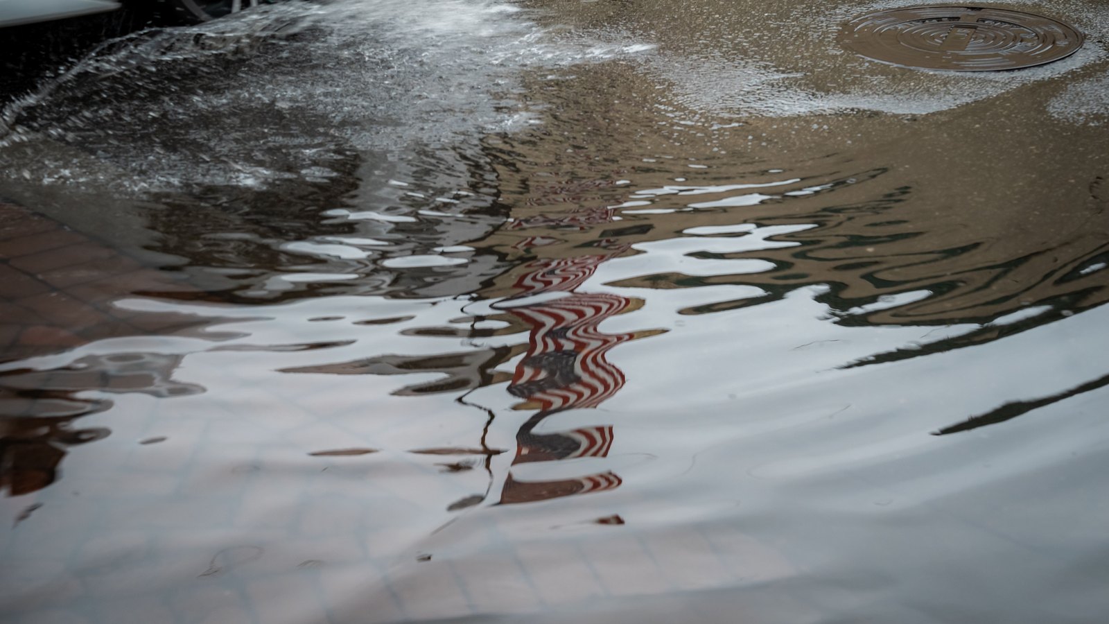 Rainwater standing across a paved street, a storm drain cover at the top right and the wheel of a parked car at the top left, with buildings reflected in the surface of the water. No person is in frame and no sign or lettering is legible. Stock photo, not a photograph of this event.