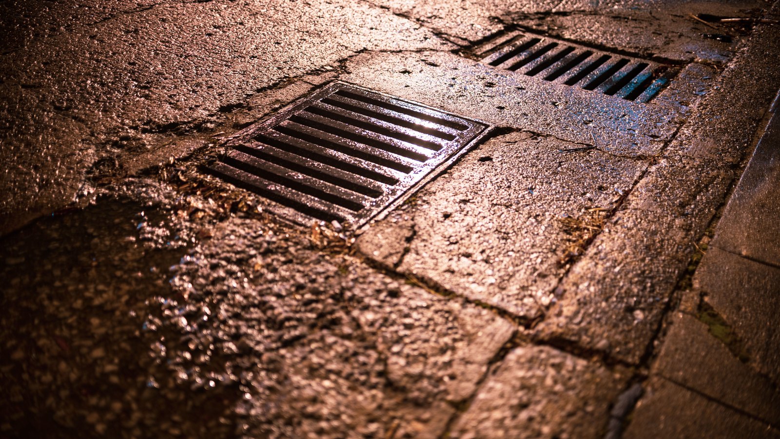 Two metal storm drain grates set into a wet, cracked paved street at night, with rainwater pooling around them and warm street lighting reflecting off the surface. No people, signage, lettering or number plates are visible in the frame. Illustrative only, not a photograph of this event.