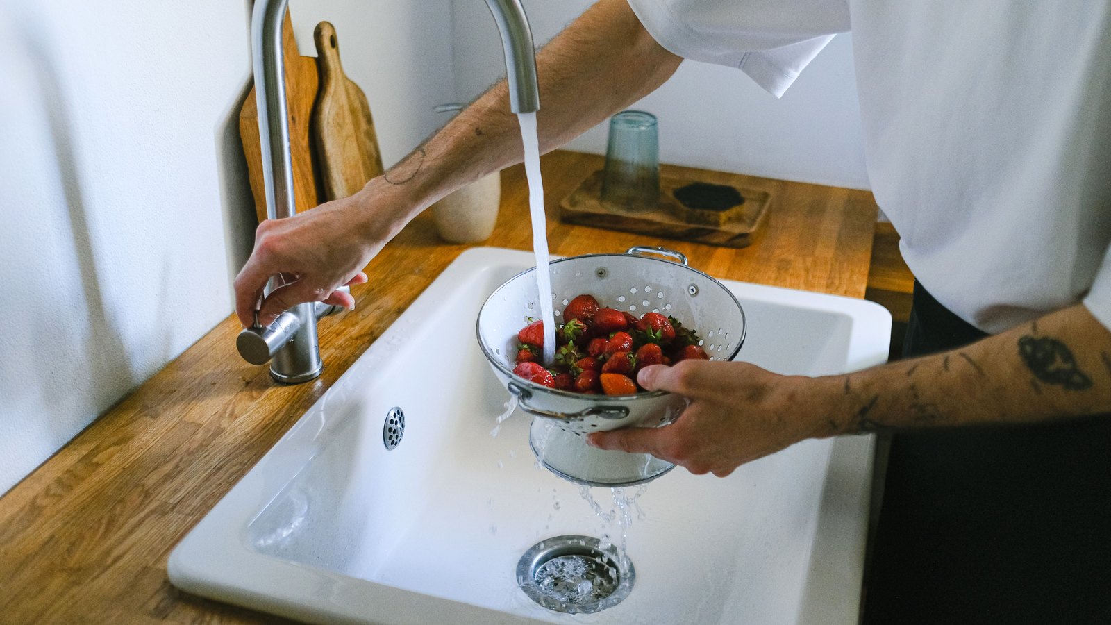 Hands rinsing strawberries beneath a running kitchen faucet, with the person cropped below the face and no legible text, not a photograph of this event.