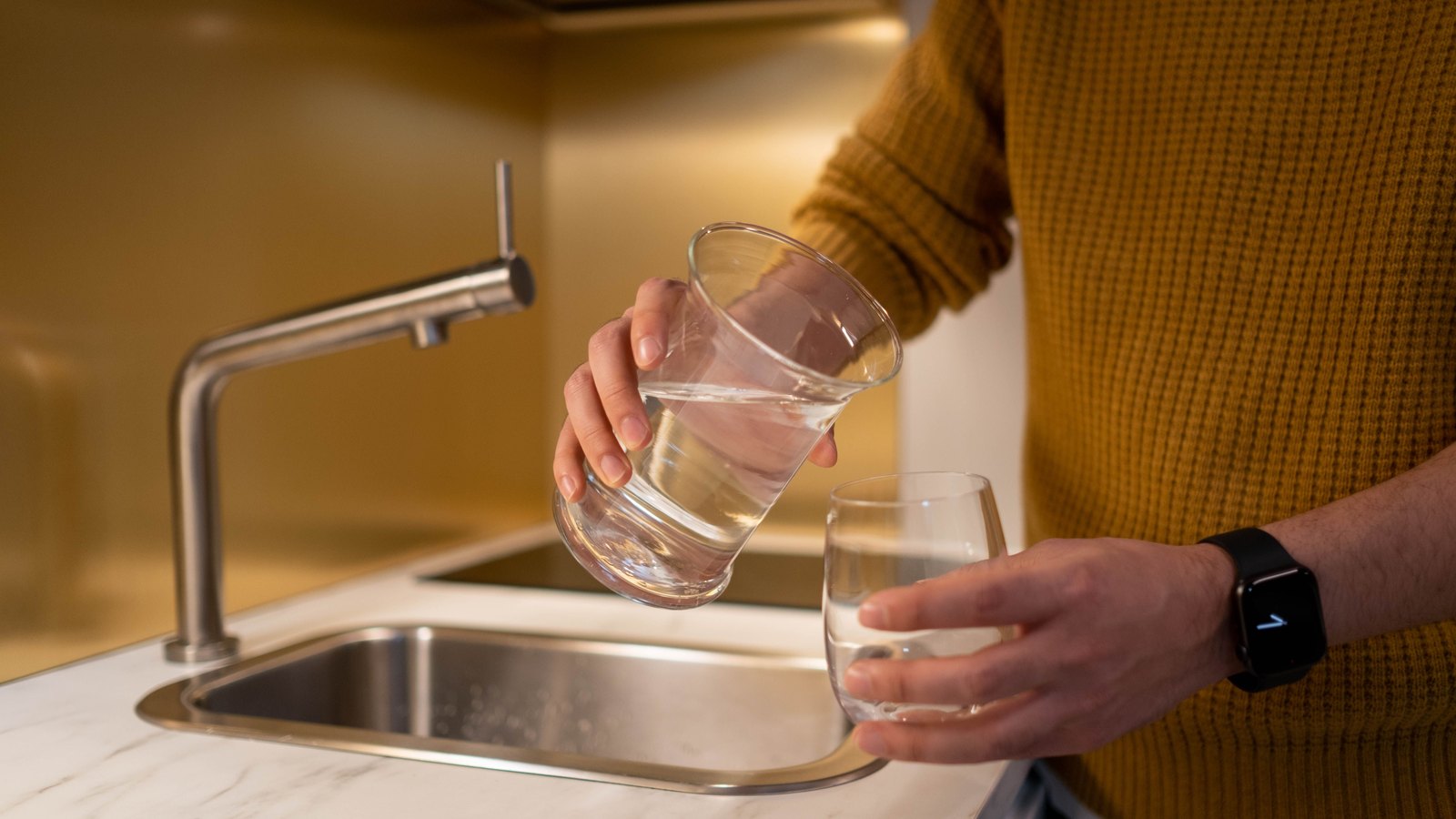A person in a mustard jumper, shown from the shoulders down, pours water from a tumbler into a second glass over a stainless steel kitchen sink, with a chrome tap to the left and no signage or legible text in view. A generic illustration, not a photograph of Cape Coral.