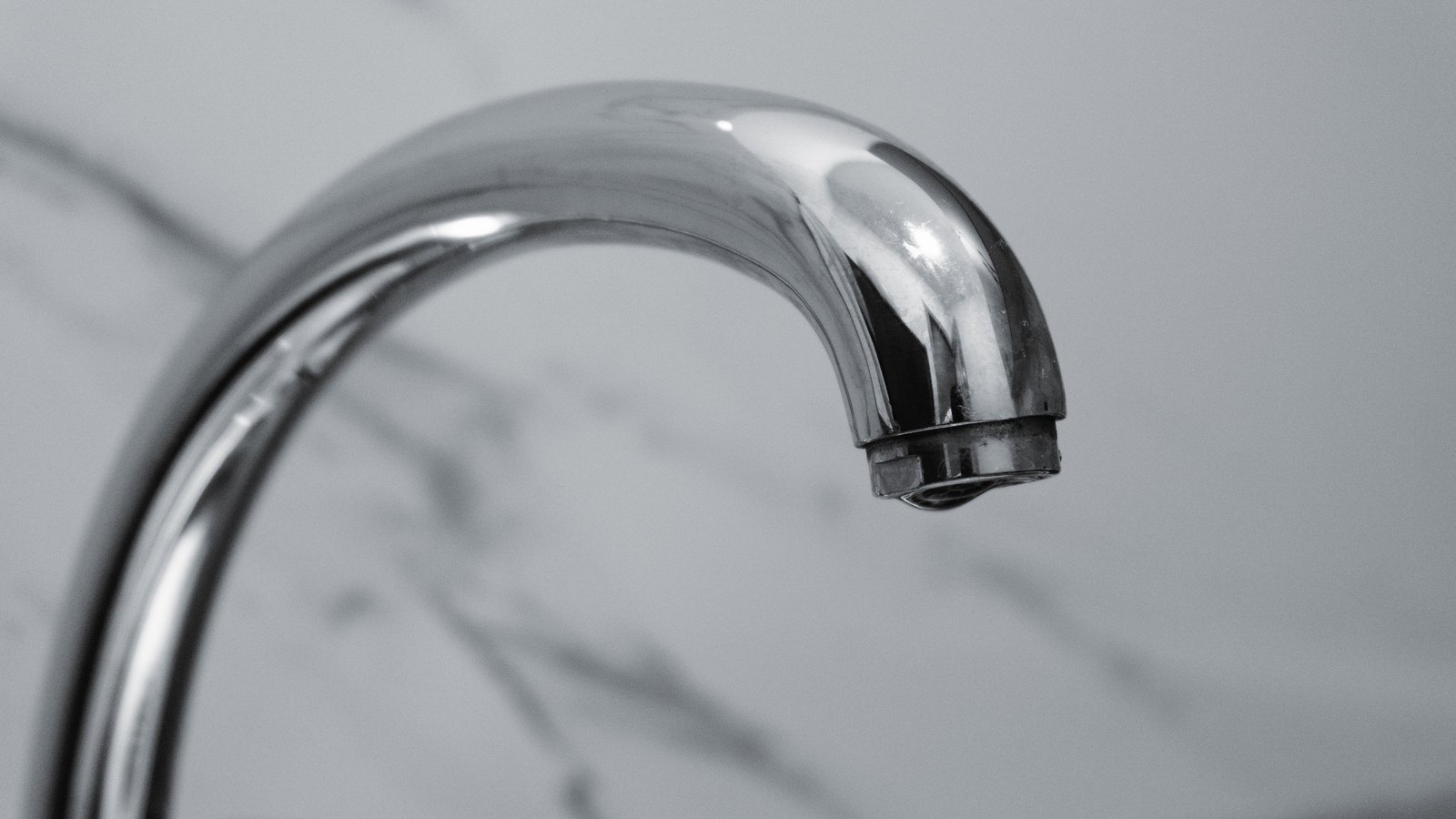 A chrome kitchen tap curving to the right against a pale marble wall, a single drop of water hanging from the lip of the spout, shot close and shallow so that nothing but the tap is in focus, with no people, signs, text or brand marks visible anywhere in the frame. A generic illustration, not a photograph of this event.