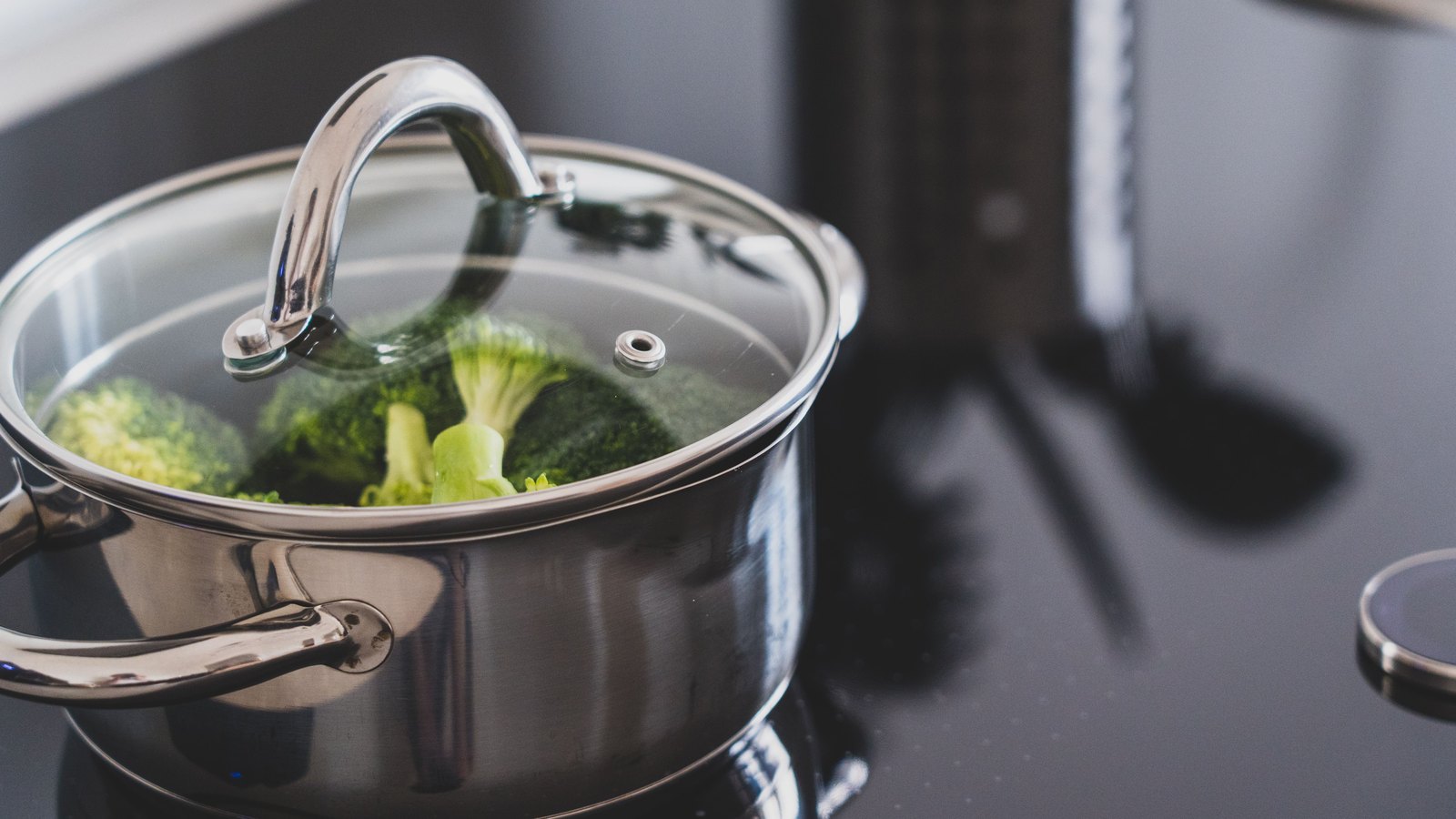 A photograph of a stainless steel saucepan with a clear glass lid sitting on a dark ceramic hob, with broccoli florets visible through the glass and condensation on the underside of the lid. A second pan handle enters at the lower left and a blurred utensil stands in the background. No people, signage, lettering or brand marks are visible and no place can be identified from it. A generic kitchen illustration, not a photograph of Cape Coral or of any address under notice.