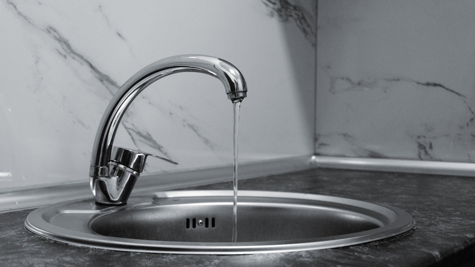 A black and white photograph of a chrome kitchen mixer tap running a thin stream of water into a round stainless steel sink set in a dark worktop, against a marble backsplash.