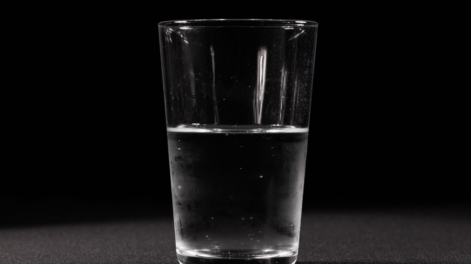 A plain drinking glass, a little over half full of still clear water, lit from the front against a black background and standing on a dark textured surface. A few small bubbles cling to the inside of the glass. There is no tap in the frame, no person, no signage, no lettering and no brand mark, and no place can be identified from it. A generic illustration of drinking water, not a photograph of Cape Coral or of any address that was under notice, and not evidence about the condition of anyone's water.