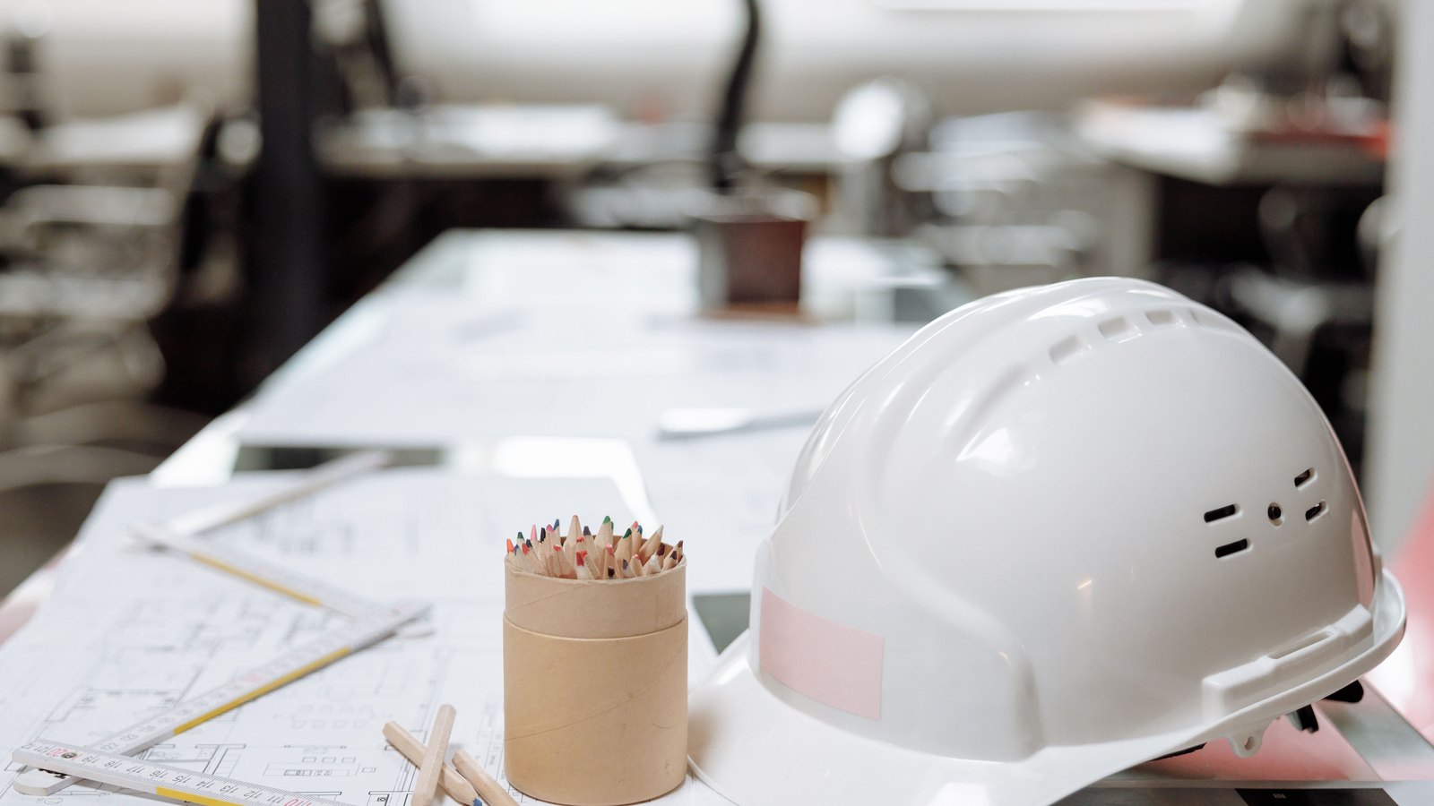 A white hard hat lying on a set of architectural floor plans on a desk, a folding wooden rule and a pot of coloured pencils beside it, an empty drawing office blurred behind. No person is in frame and no text on the drawings is legible. Stock photo, not a photograph of this event.