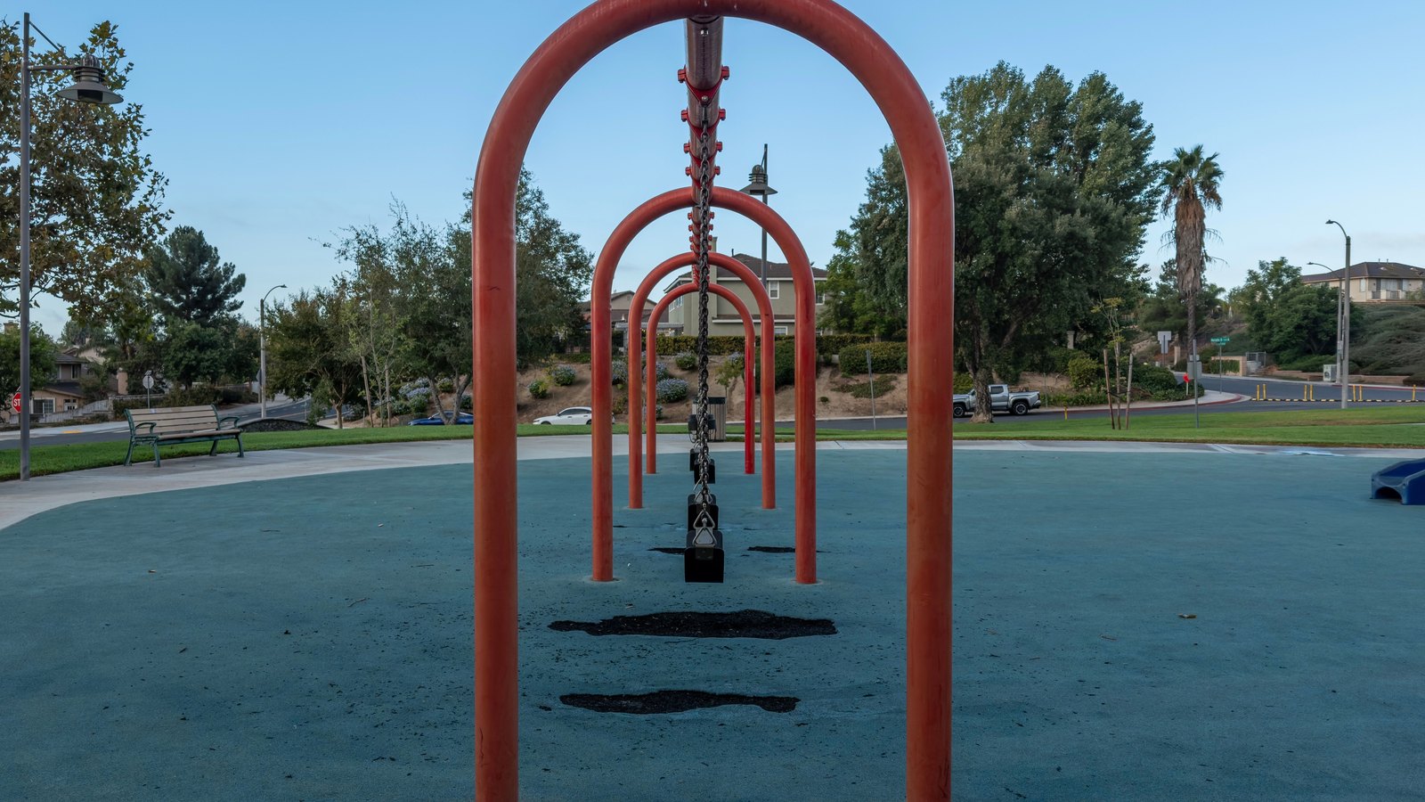 An empty row of swings hanging from red steel arches stands on blue rubber safety surfacing in a public park at dusk, with unlit lamp standards, a bench, palms and other trees behind it and houses on a low rise beyond. A distant street sign at the roadside is too small to read, and no person, agency marking or identifiable place is visible in the frame. It is a stock photograph and not a photograph of this event.
