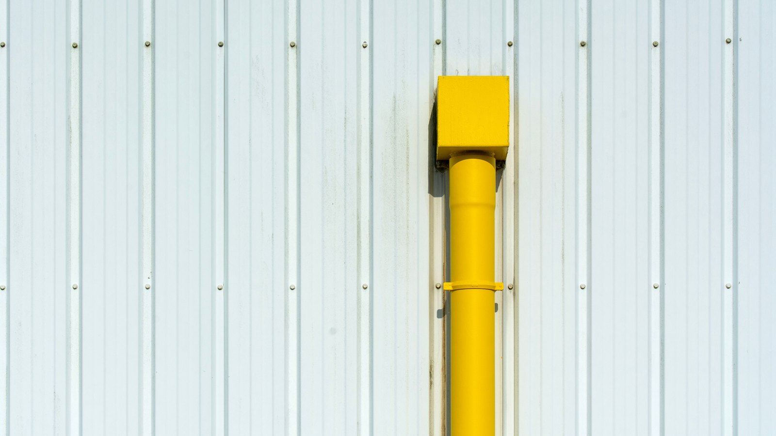 A yellow painted pipe running vertically down the face of a white corrugated metal wall, emerging from a plain yellow box near the top and held by a single bracket. The frame carries no text, no signage, no logo, no person and nothing that identifies a place. It is a stock photograph of a pipe on a wall, not a photograph of West Cape Estates or of any Cape Coral utilities work.