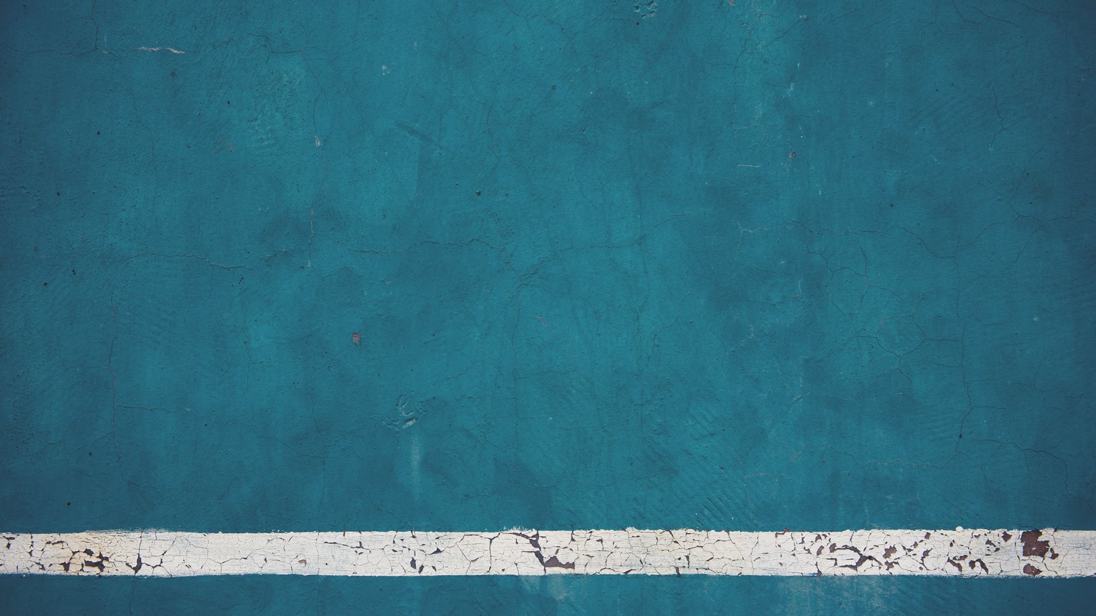 A flat teal painted wall filling the frame, its surface covered in a fine web of hairline cracks, with a white painted horizontal stripe across the bottom where the paint has flaked away to bare patches. No person, no window, no signage and nothing identifying a place appears. A generic photograph of a cracked painted surface, not a picture of any home in this story.