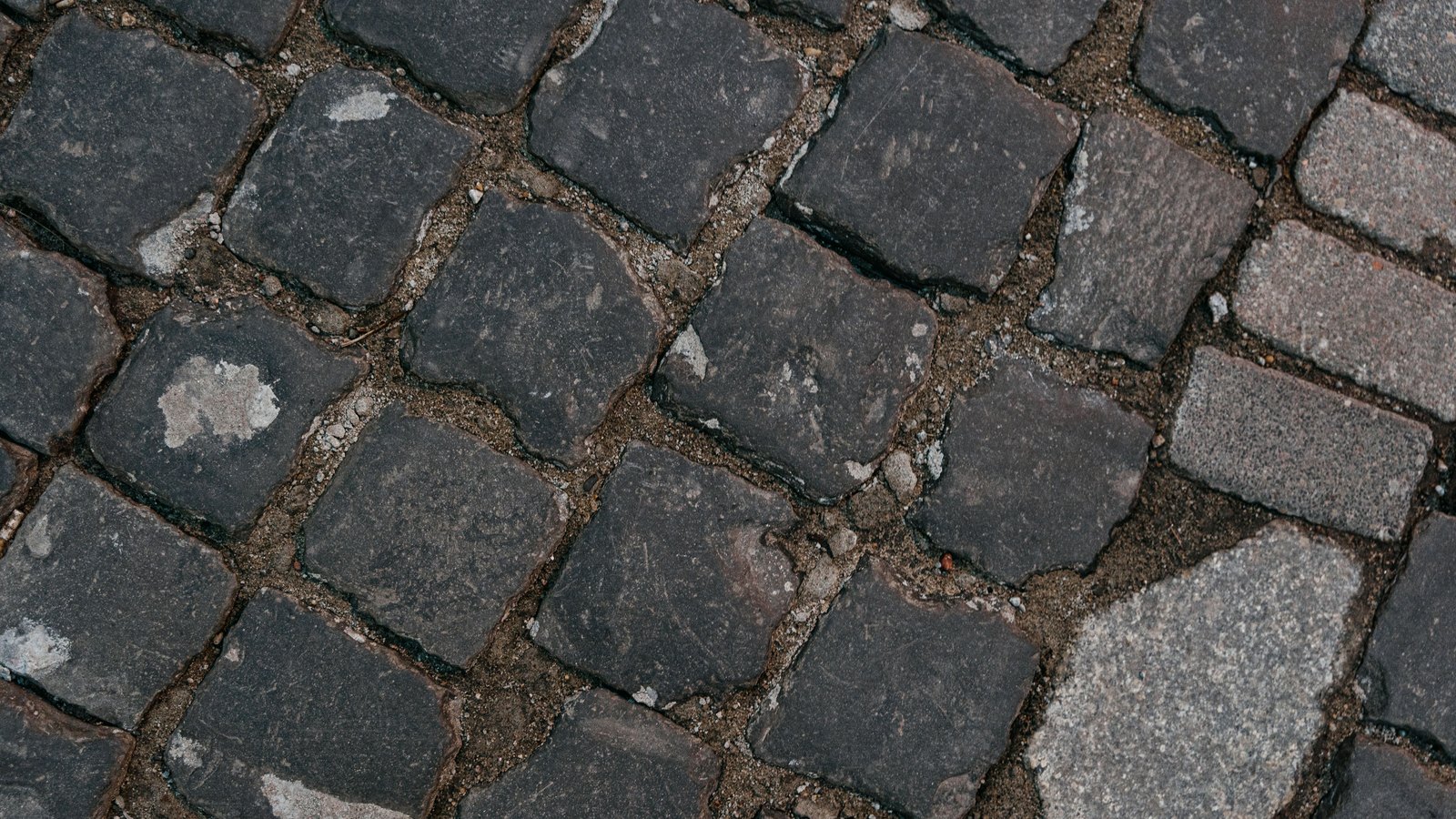 Dark grey stone setts fill the whole frame, seen from directly above and running on the diagonal, each block worn smooth on top with grit and pale chippings packed into the joints. A band of lighter granite blocks crosses the lower right corner. No horizon, kerb, vehicle, person or lettering is in the picture.