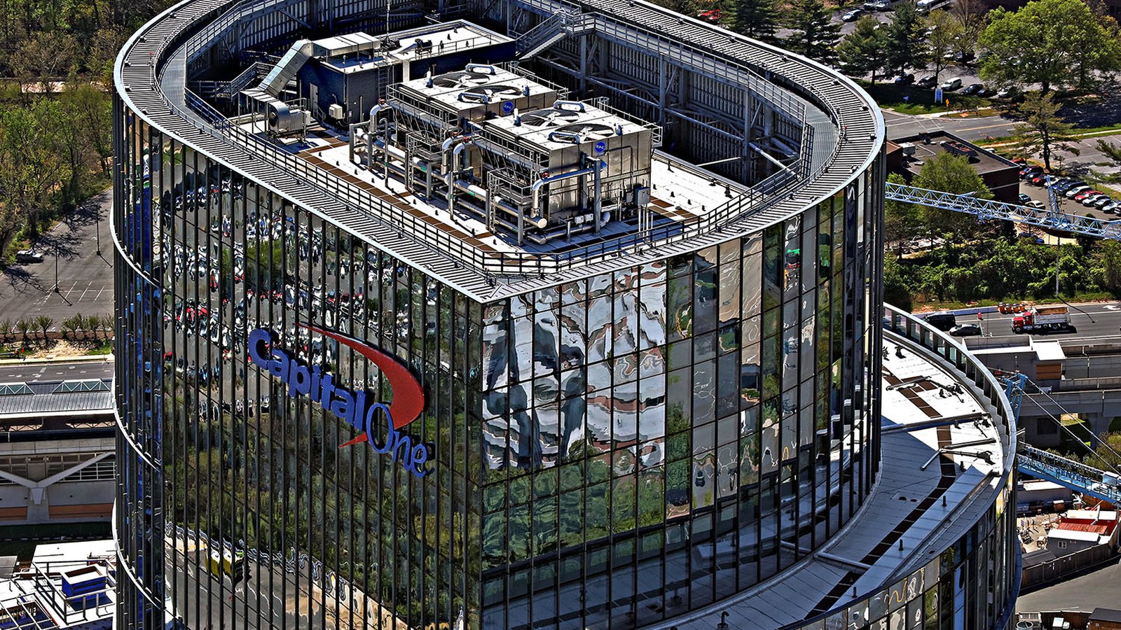 The upper floors of the Capital One Tower, the company corporate headquarters at Tysons in Virginia, photographed from the air on a clear day. The company wordmark sits on the dark glass curtain wall, with rooftop plant behind a curved parapet above it, and roads, car parks, trees and a tower crane on the surrounding development below.