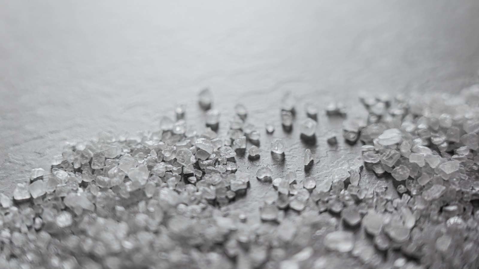 A close photograph of coarse translucent salt crystals scattered in a loose band across a pale grey surface, sharp in the middle of the frame and falling out of focus towards the top.
