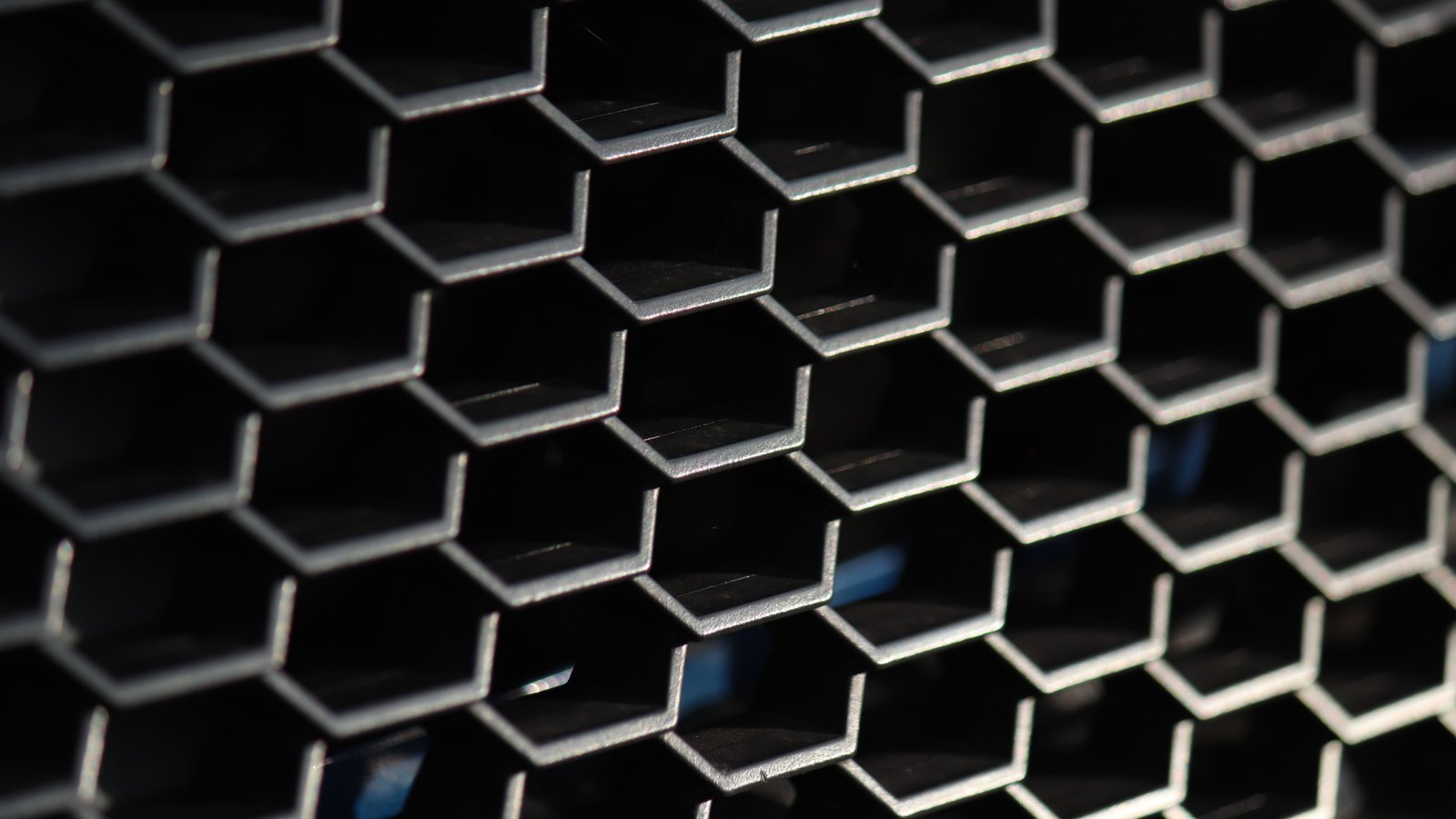A close view of a honeycomb car grille, rows of hexagonal metal cells angling away into shadow with a faint blue reflection behind them.