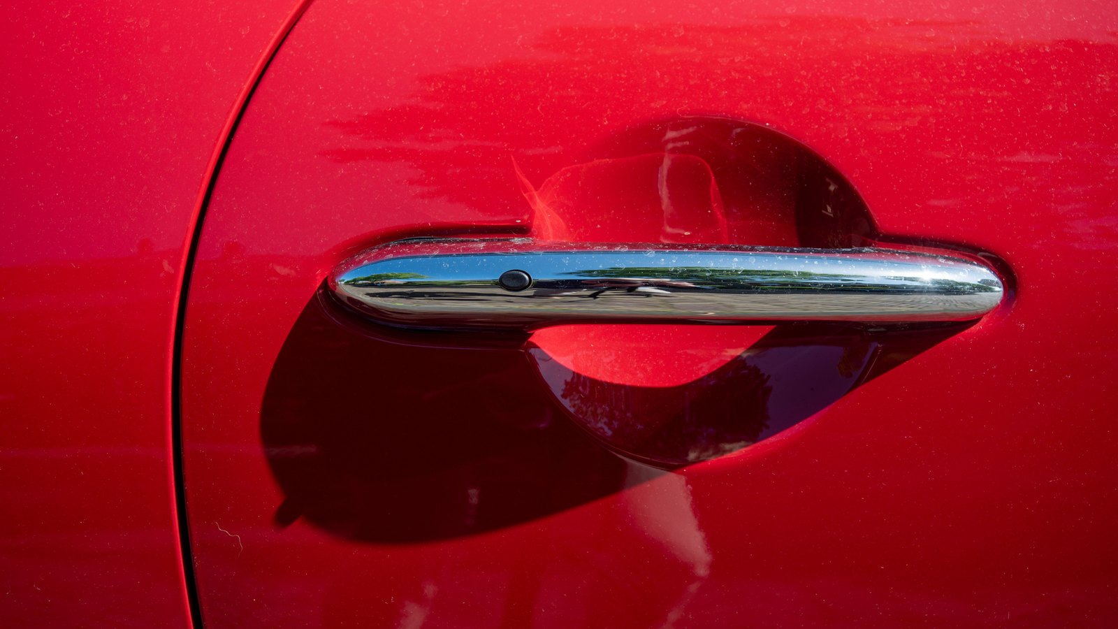 A chrome door handle set into the glossy deep red paintwork of a car door, photographed close up in daylight with the surrounding trees showing only as a faint reflection in the chrome. No badge, no lettering, no registration plate, no place and no person appears in the frame.