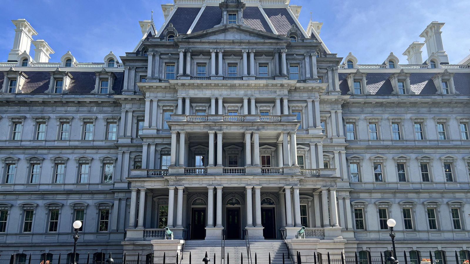 The Eisenhower Executive Office Building in Washington, its grey granite front rising in tiers of paired columns under steep slate mansard roofs, with a flight of steps and two bronze lamp standards at the central entrance.