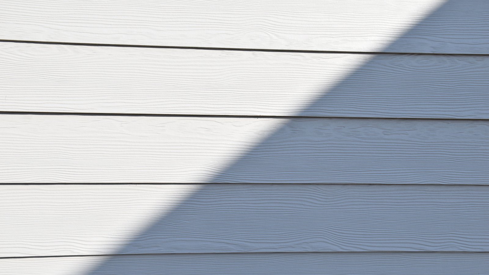 White painted lap siding filling the frame, the grain of each board just visible, with the diagonal shadow of a roofline falling across the right half.