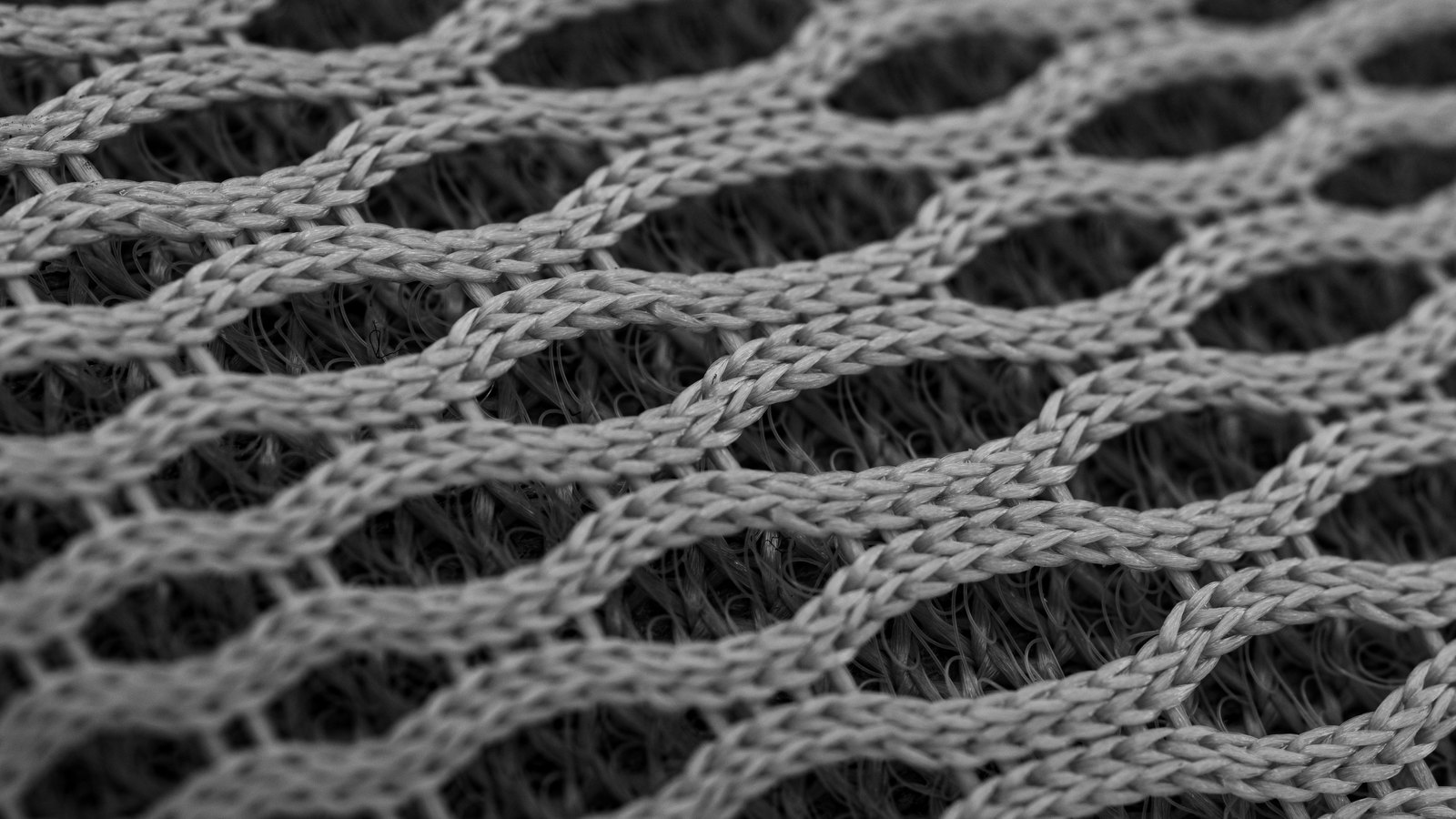 A macro view of grey knitted nylon mesh, braided cords crossing in a regular diamond lattice with soft shadow showing through the openings.