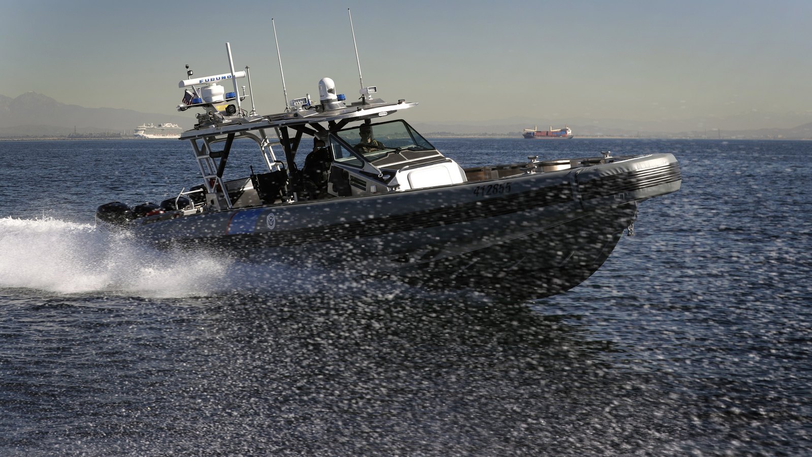 A United States Customs and Border Protection Air and Marine Operations 41-foot Coastal Interceptor running at speed across open coastal water, throwing spray, with radar and antenna masts above the console and distant ships on the horizon.