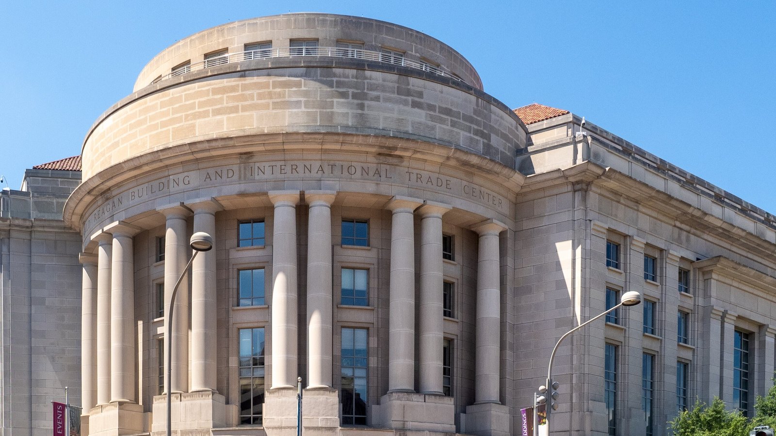 The limestone rotunda of the Ronald Reagan Building in Washington, which houses Customs and Border Protection headquarters, its colonnade under a carved band reading Reagan Building and International Trade Center against a clear blue sky.