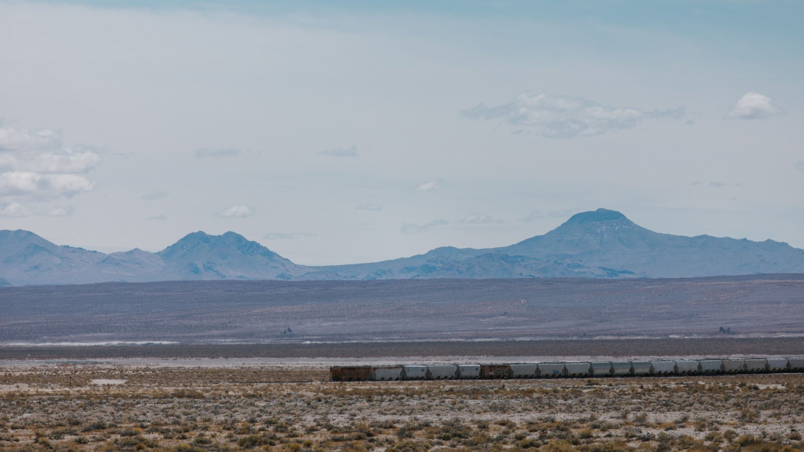 A long freight train of pale covered hopper cars crossing a wide arid basin in the American West, seen from a distance across sagebrush flats, with low bare mountain ranges on the horizon under a pale sky. No lettering, marking or person is legible at this range.