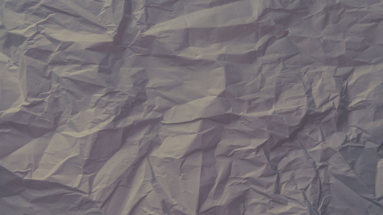 A sheet of pale paper that has been crumpled and flattened out again, photographed square on and filling the frame. Creases run in every direction, catching the light along their folds and leaving soft grey shadows between them. The paper is blank, with no printing, writing or markings of any kind.