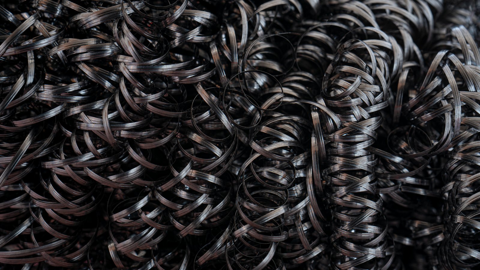 A tight overhead view of dark steel machining swarf, hundreds of tightly curled metal spirals filling the frame.