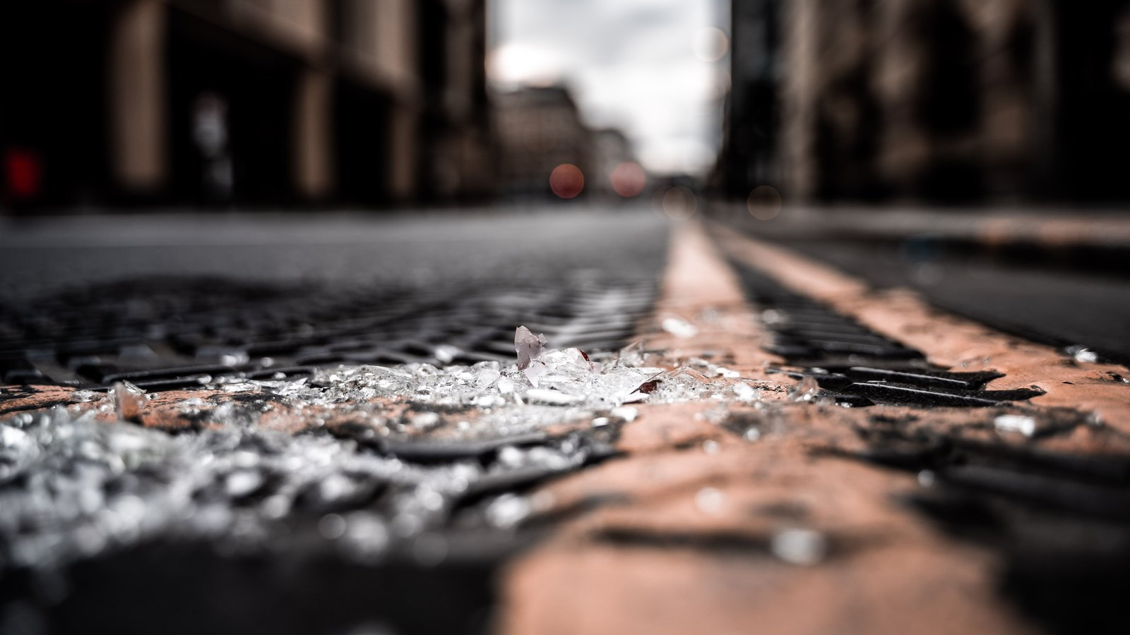 Shards of broken vehicle safety glass lie scattered along a kerb and across a drain grate on a street, with the roadway and buildings behind thrown completely out of focus. No person, vehicle, number plate, agency marking or identifiable place is visible in the frame. It is a stock photograph and not a photograph of this event.