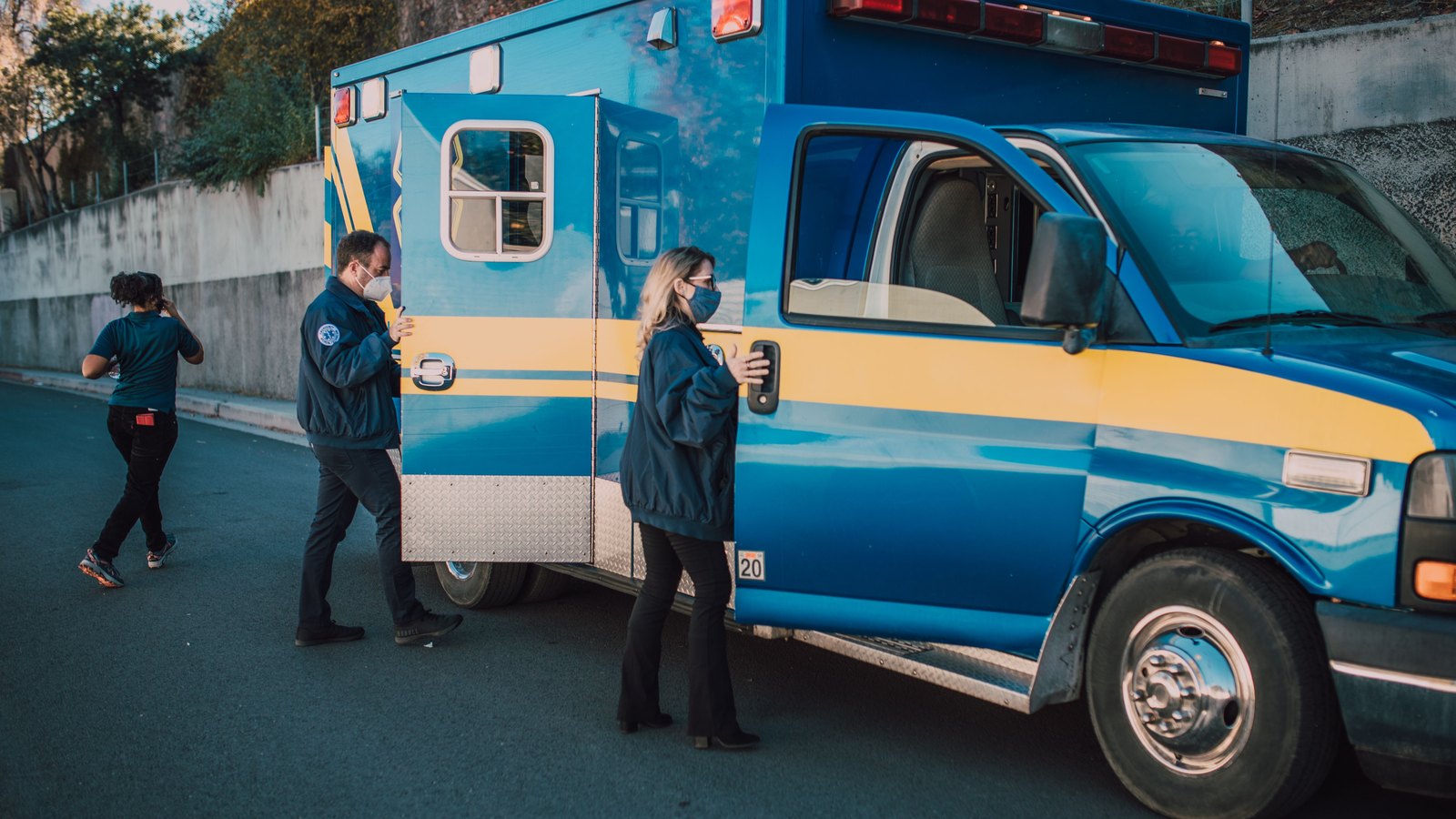 Three emergency medical workers in dark uniforms and face masks move around the open rear doors of a blue and yellow ambulance parked on a paved road beside a concrete retaining wall, with green trees and daylight behind it. No agency name, unit lettering or number plate is legible in the frame. It is a stock photograph and not a photograph of this event.