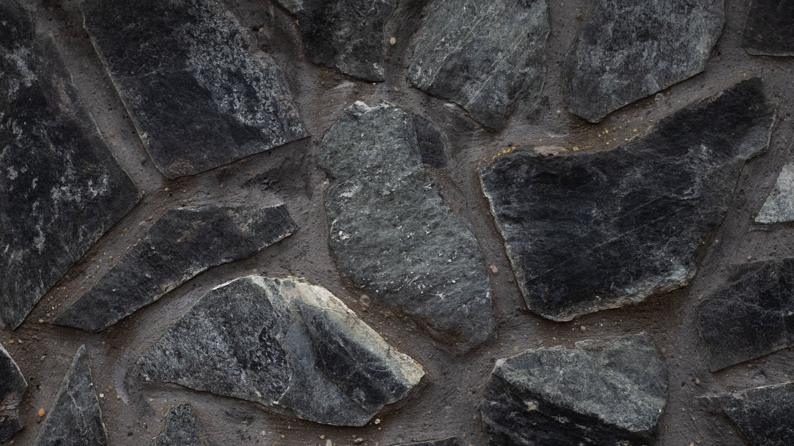 Dark grey and near black stone slabs of irregular shape set into pale grey mortar, filling the frame as an intact flat wall surface, with no text, people or landmarks in view.
