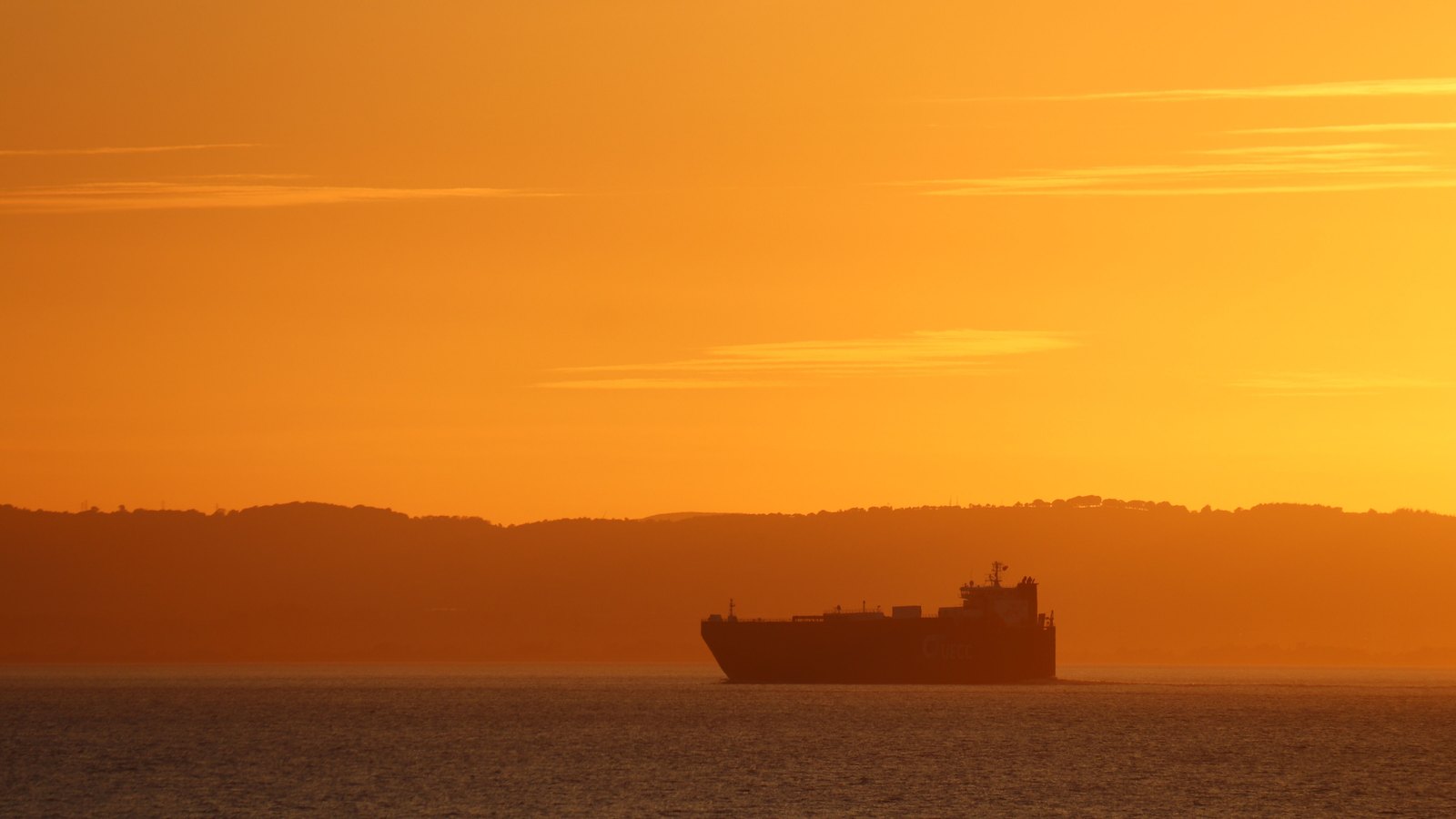 A cargo vessel in silhouette on calm water at sunset, with a low headland behind it and an orange sky above.