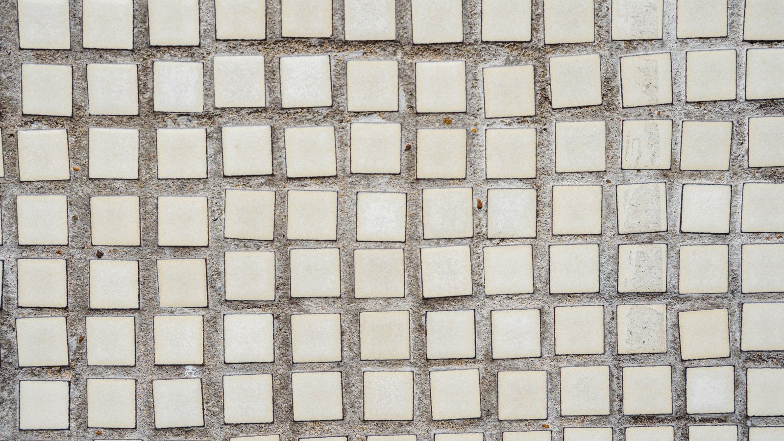 A flat overhead view of small pale square ceramic tiles set in coarse grey grout, row upon row of near-identical units filling the frame.