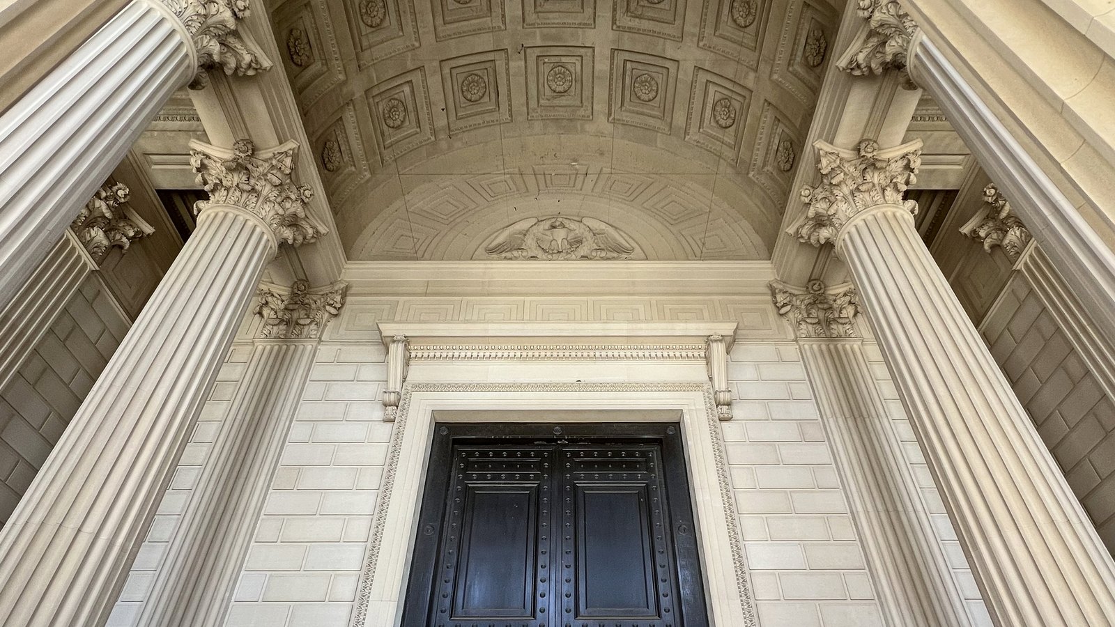 Looking up into the portico of the National Archives Building in Washington, between two fluted Corinthian columns to a coffered stone vault, a carved eagle set above the door frame, and the upper part of a pair of tall dark bronze doors below.