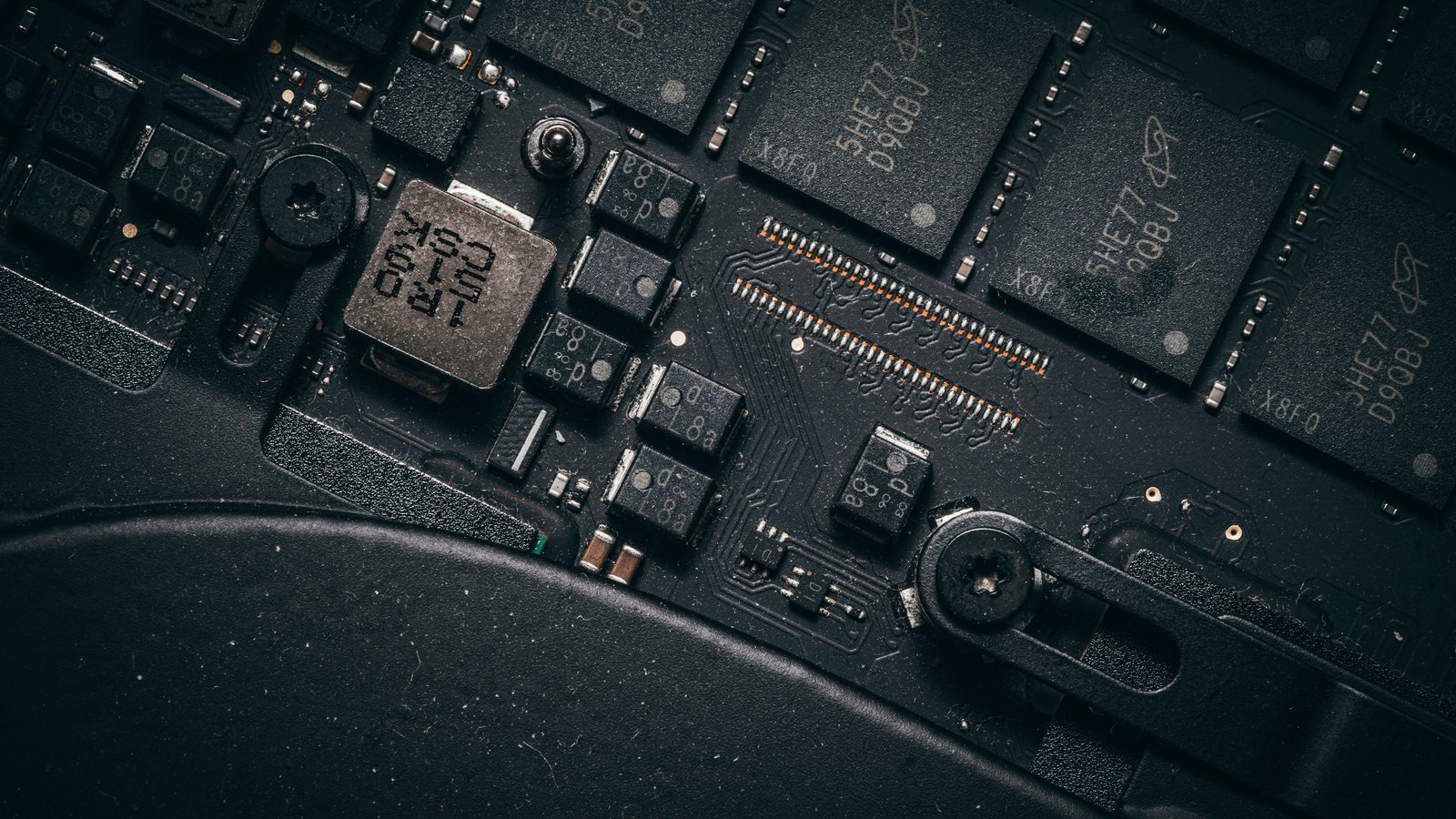 A close-up of a dark circuit board, showing rectangular memory chips, small square surface-mounted components and a row of fine metal contacts, lit from one side.
