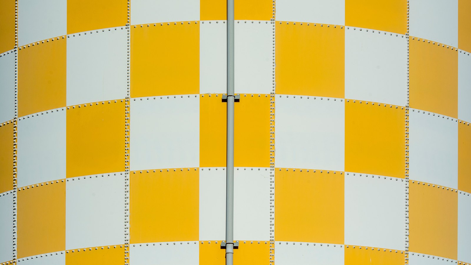 A curved riveted wall painted in a checkerboard of yellow and white panels, rows of rivet heads marking the seams, with a slim vertical pipe running down the centre of the frame.