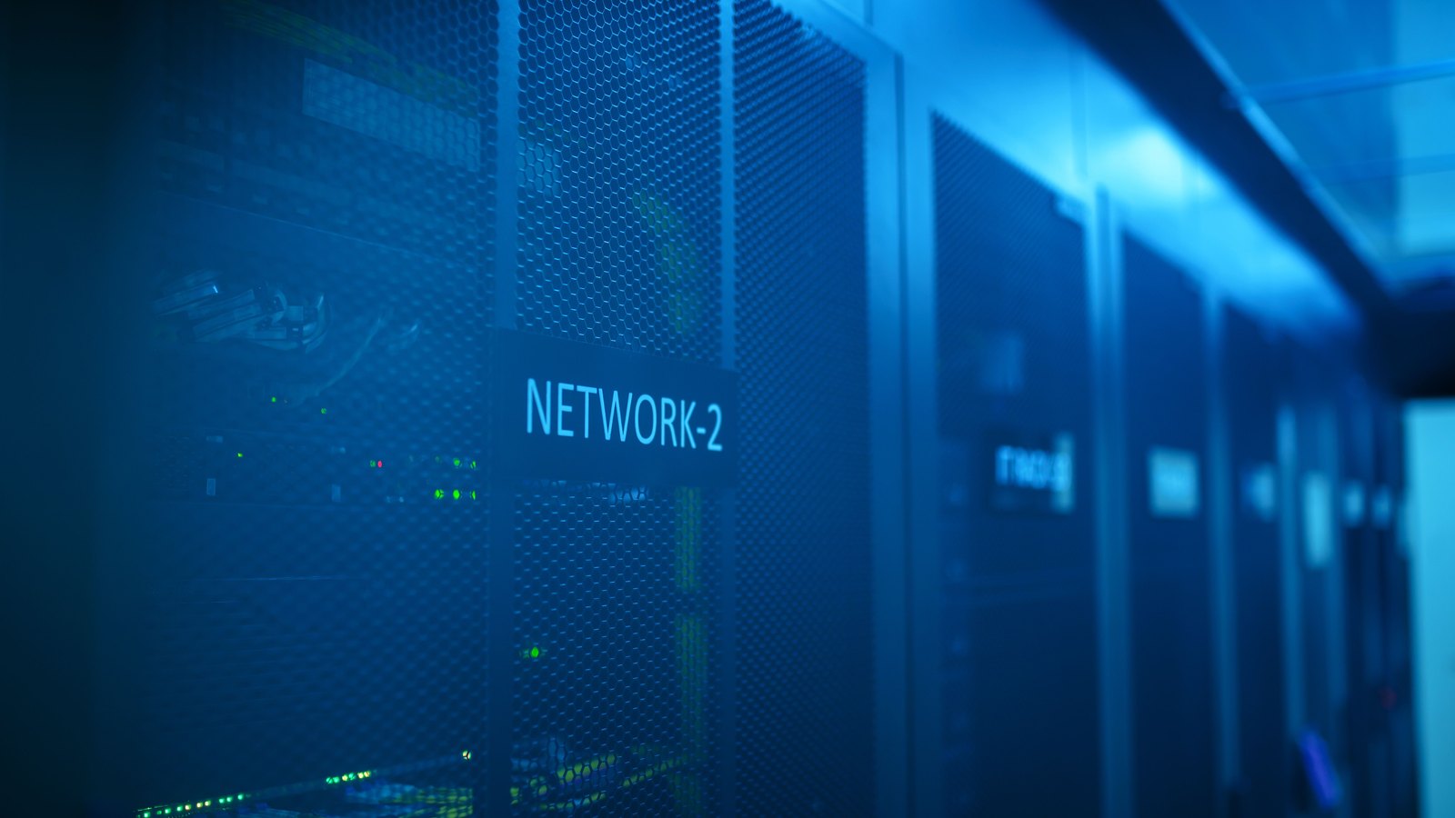 A dim aisle in a data hall lit blue, with tall perforated server cabinet doors receding into the distance and small green status lights showing through the mesh. An illustrative photograph, not a picture of any facility named in the request for comment.