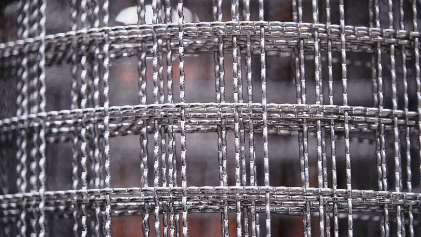 A close-up of bright woven steel wire mesh, the wires crossing in a regular square grid, with the background thrown completely out of focus behind it.