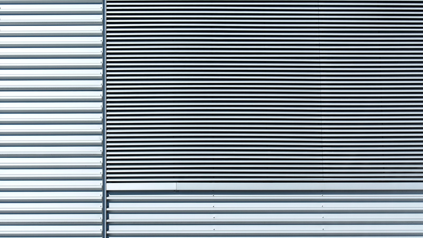 A pale grey aluminium building wall photographed square on. Vertical ribbed cladding runs down the left side, a wide band of close set horizontal louvre slats fills the centre, and further horizontal ribbed panels run below. No sky, ground, sign or person is in the frame.