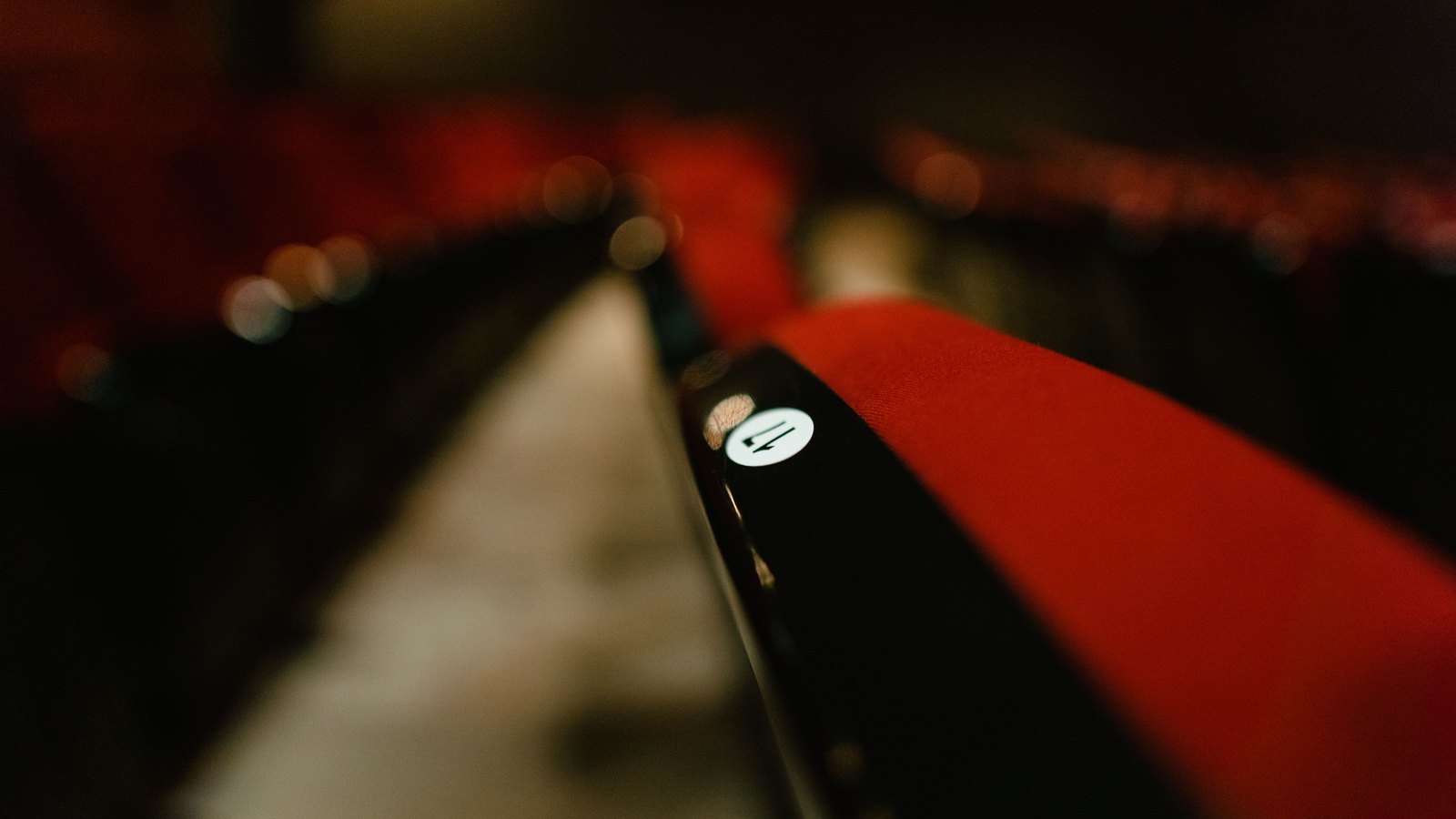 A close photograph of the end of an empty row of red upholstered seats in a darkened auditorium, the black armrest carrying a small white disc with a single seat number on it, further rows falling away into soft focus behind.